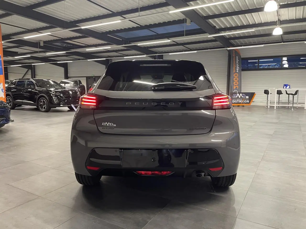 Vue arrière d'une Peugeot 208 hybride gris selenium avec feux arrière LED 3 griffes et bandeau noir brillant en intérieur showroom.