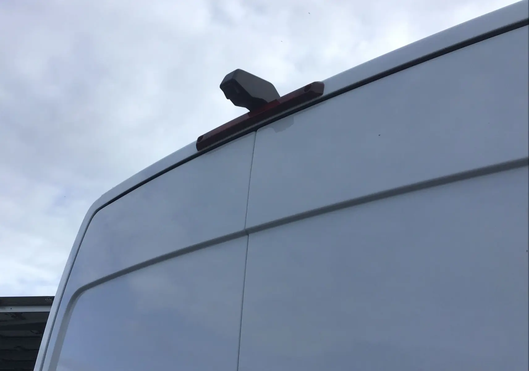 Gros plan sur la caméra de recul installée en hauteur à l'arrière d'un Renault Master blanc, vue en légère contre-plongée.