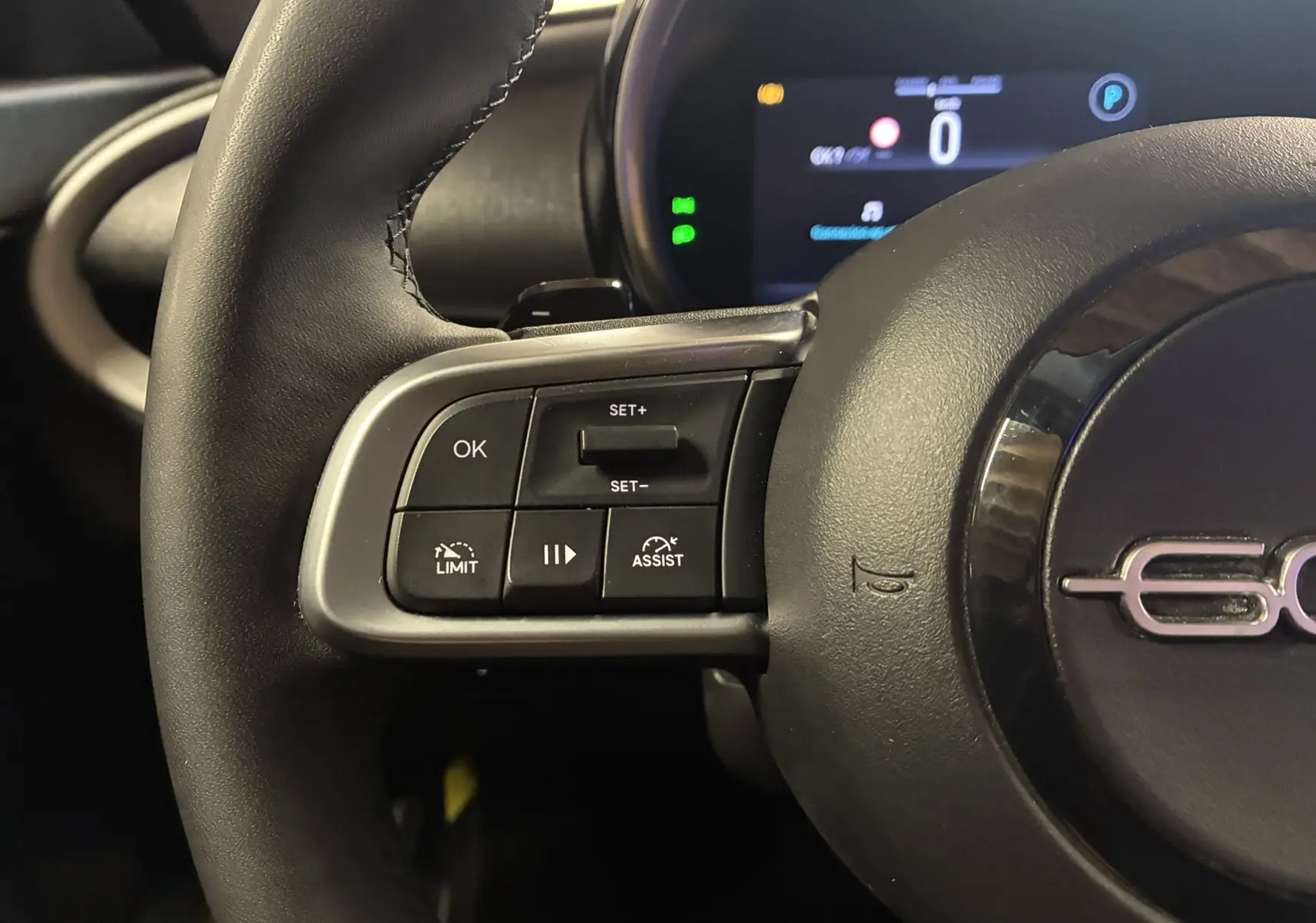 Gros plan sur le volant noir de la Fiat 600 1.2 Hybrid 2024, avec commandes intégrées et tableau de bord numérique en arrière-plan.