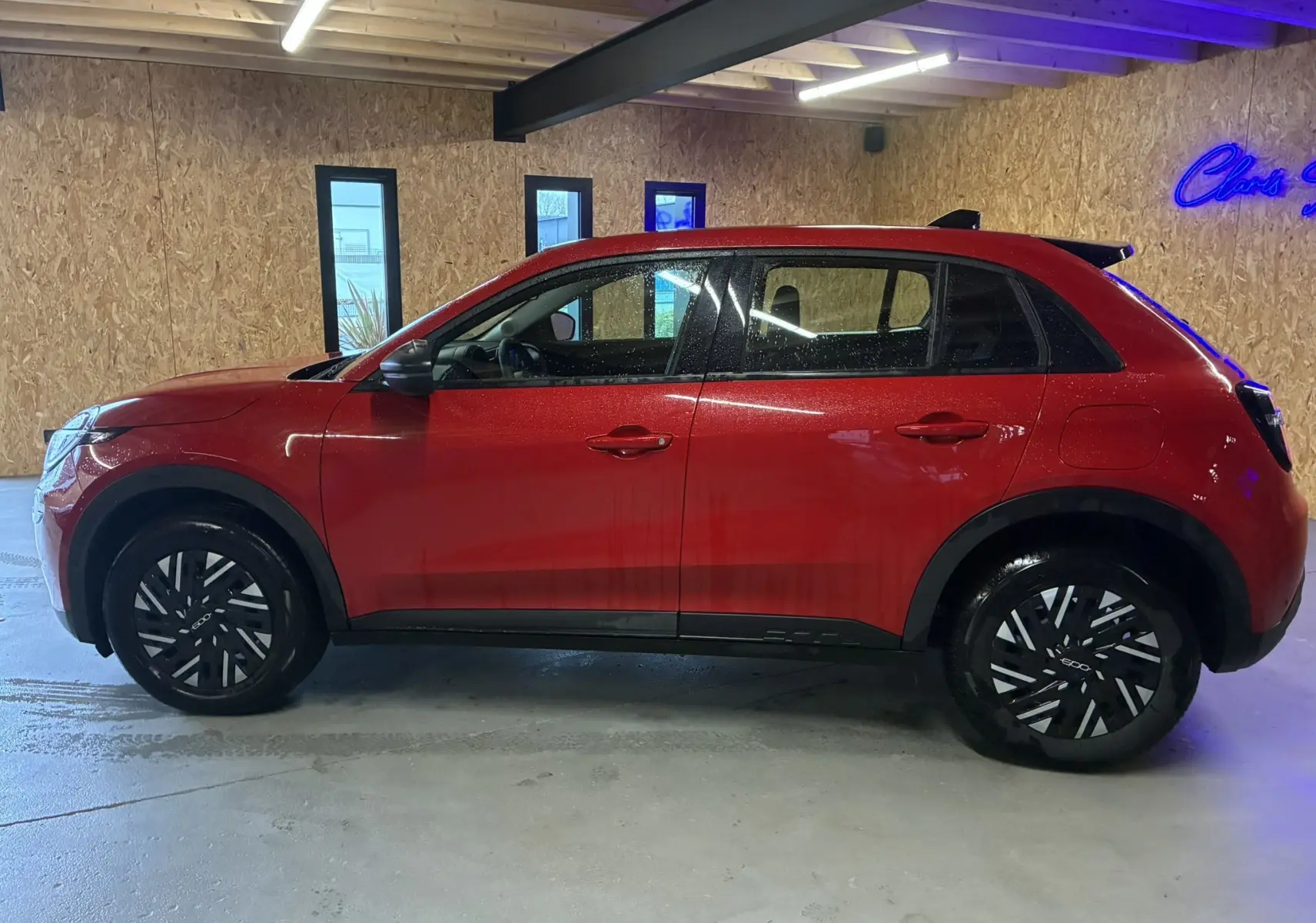Vue latérale droite d'une FIAT 600 1.2 Hybrid 2024 rouge, avec jantes noires et toit noir contrasté dans un garage.