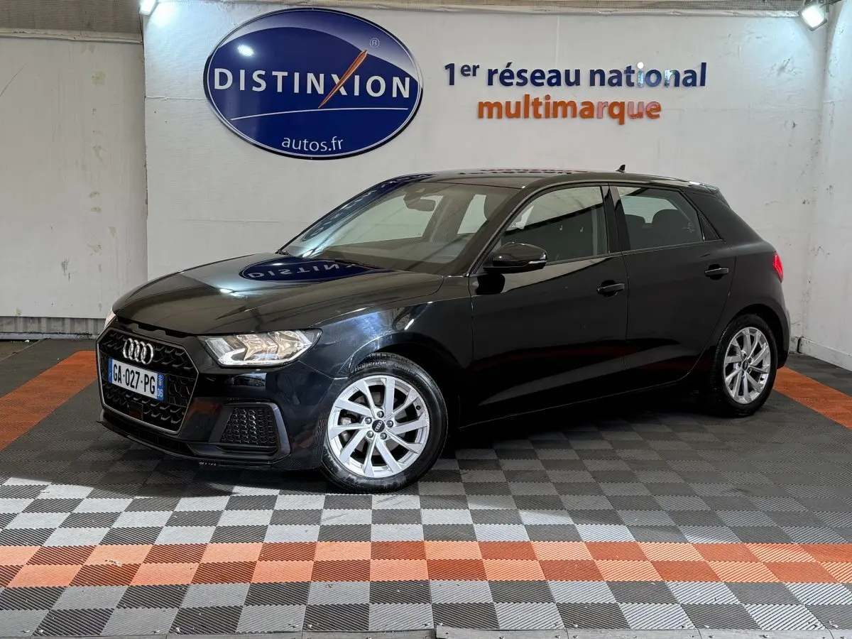 Audi A1 noire vue en 3/4 avant droit dans un showroom avec sol à damiers gris et orange.