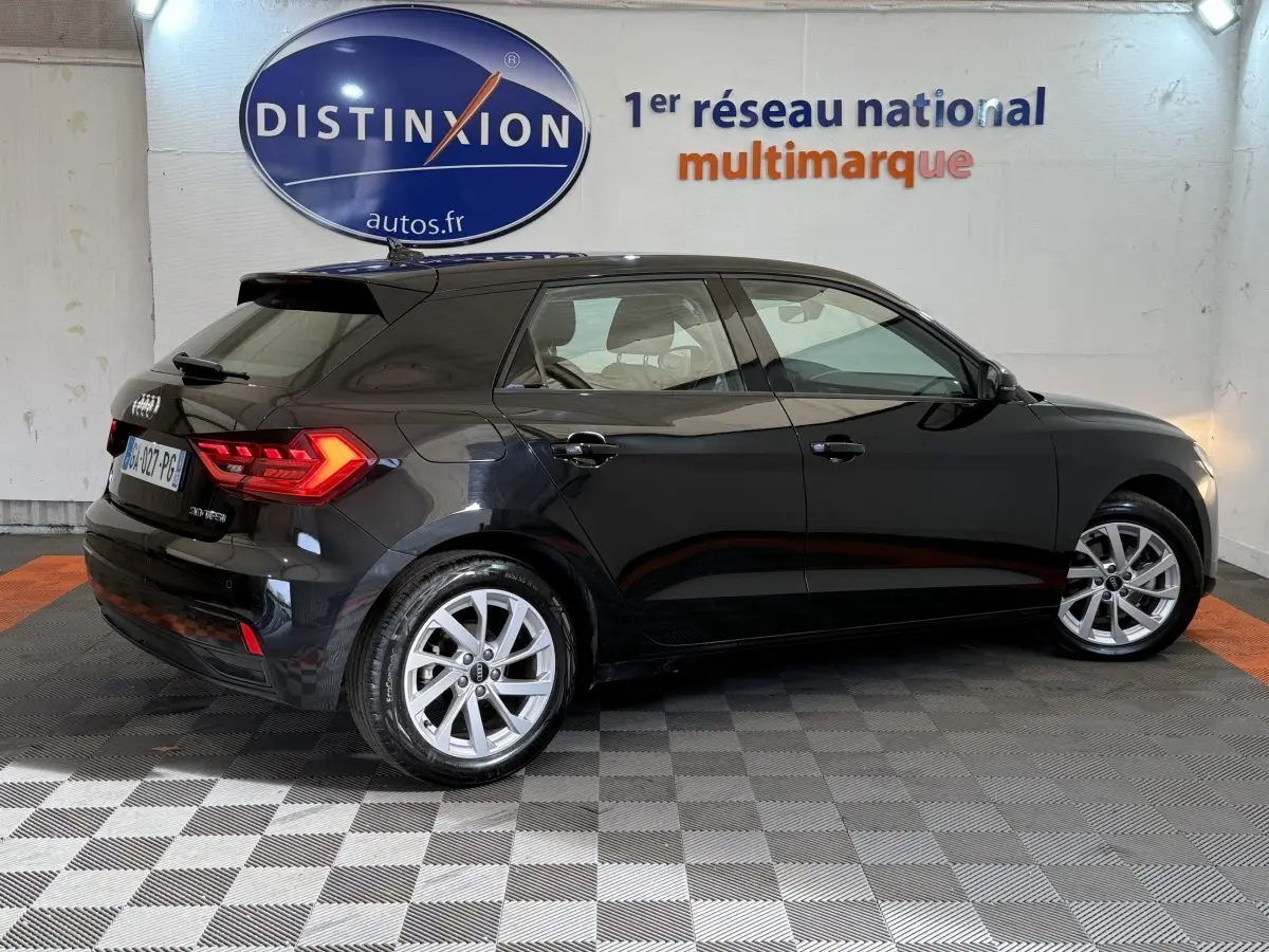 Audi A1 noire vue de profil côté droit, avec jantes argentées et feux arrière allumés en intérieur showroom.