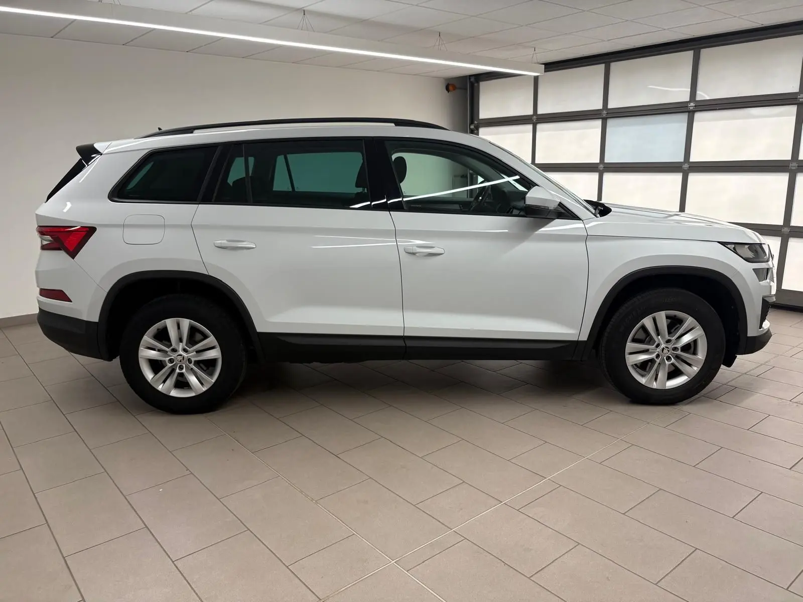 Vue de profil côté gauche d'un Skoda Kodiaq blanc Lune 2022 en intérieur, avec jantes alliage et attelage escamotable.