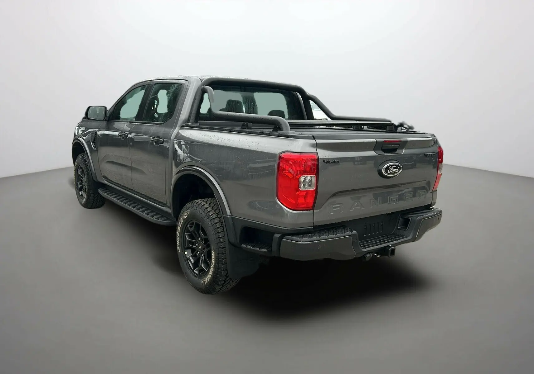 Vue 3/4 arrière droite du Ford Ranger gris carbone avec arceaux noirs et attelage visible sur fond neutre.