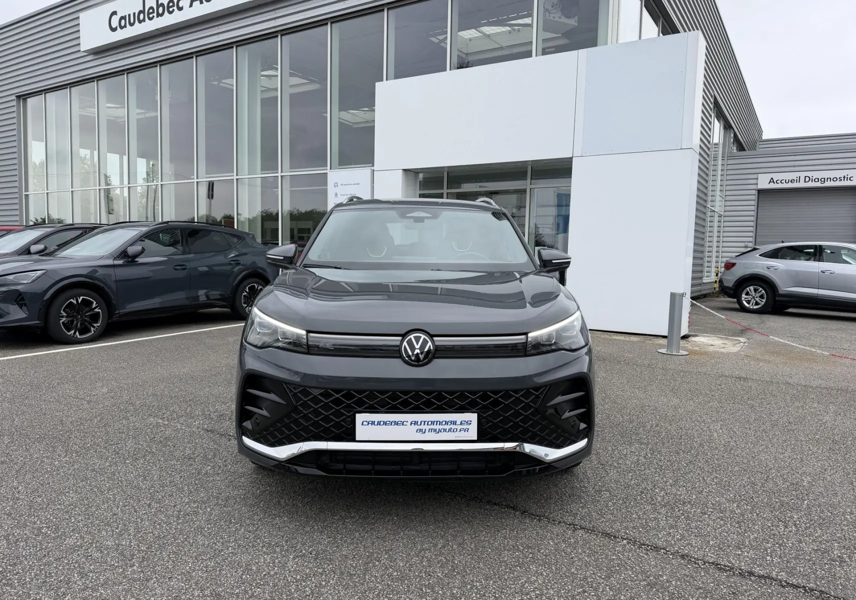 Vue avant d'un Volkswagen Tiguan gris 2025 avec calandre noire et logo VW centré, stationné devant un showroom.
