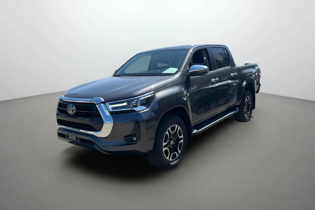 Toyota Hilux 4x4 gris atlas en 3/4 avant droit, avec jantes alu 18 pouces et marchepieds chromés visibles.