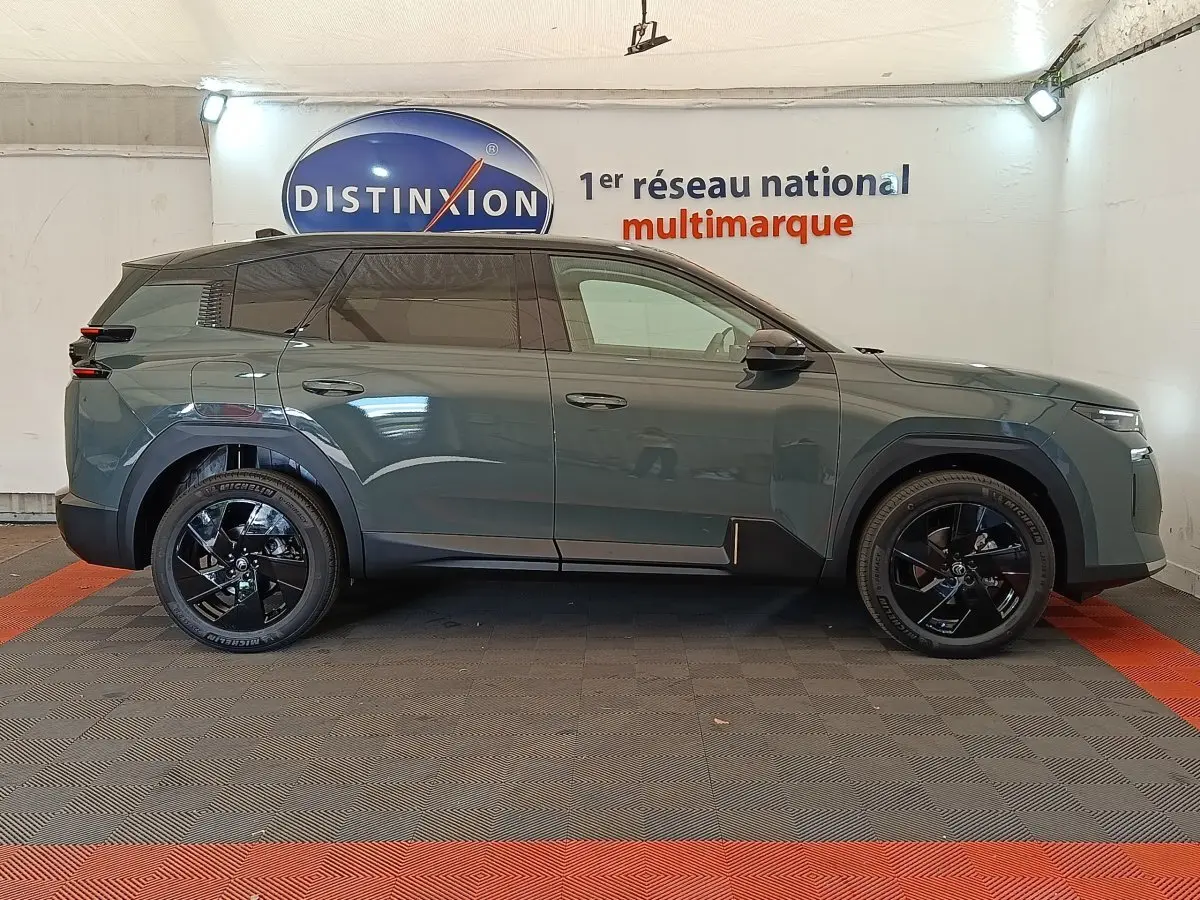 Vue de profil côté gauche d'une Citroën C5 hybride vert foncé avec jantes noires et toit noir en intérieur showroom.