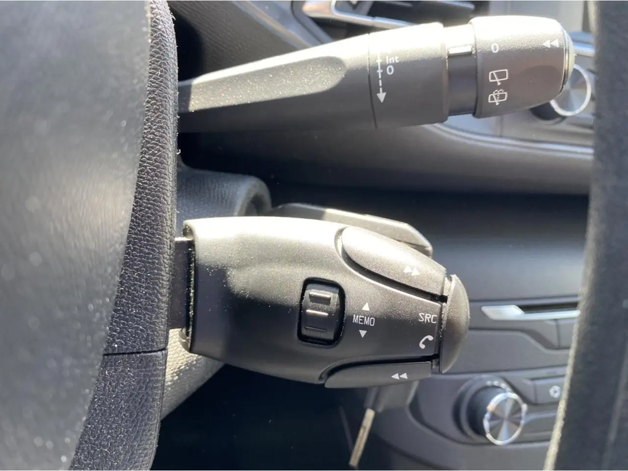 Gros plan sur la commande multifonction noire côté gauche du volant d'une Peugeot 308 SW gris clair, avec tableau de bord en arrière-plan.