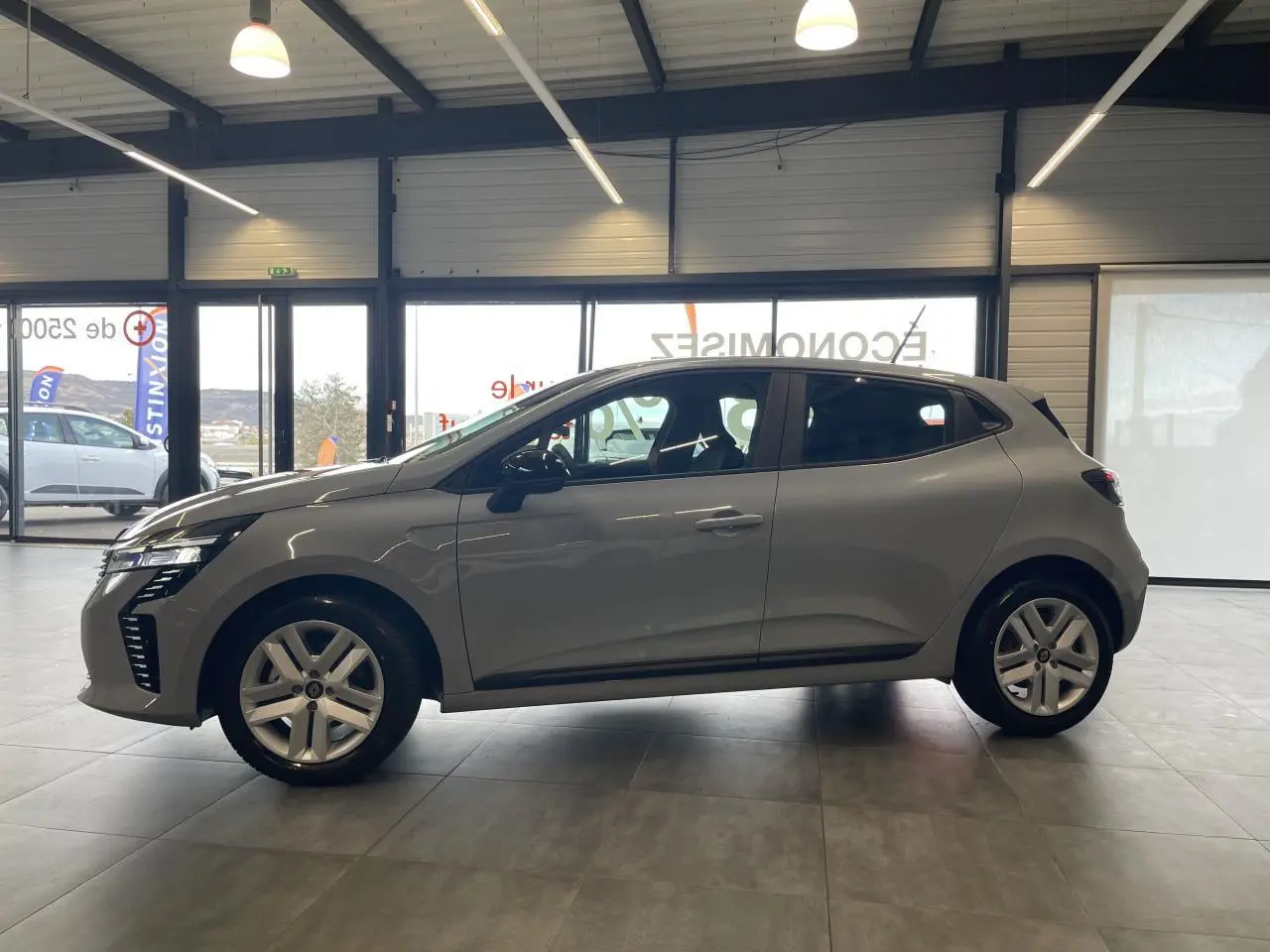 Profil côté gauche d'une Renault Clio 1.5 Blue dCi 2025 gris Rafale en showroom avec jantes alliage et poignées couleur carrosserie.