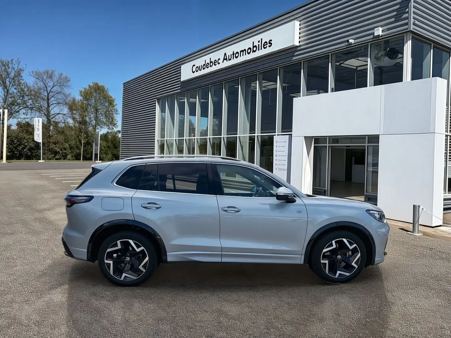 Vue latérale droite du Volkswagen Tiguan gris 2025 avec jantes alliage noires et toit ouvrant, devant un bâtiment moderne.