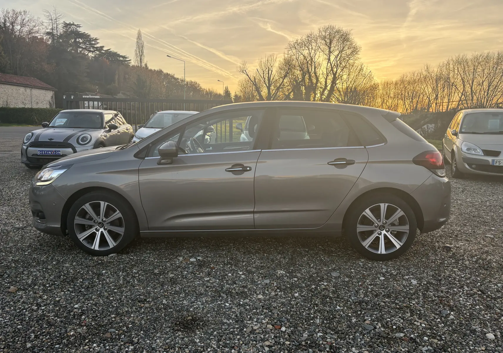 Profil côté gauche d'une Citroën C4 II gris métallisé de 2015, stationnée sur un sol gravillonné au coucher du soleil.