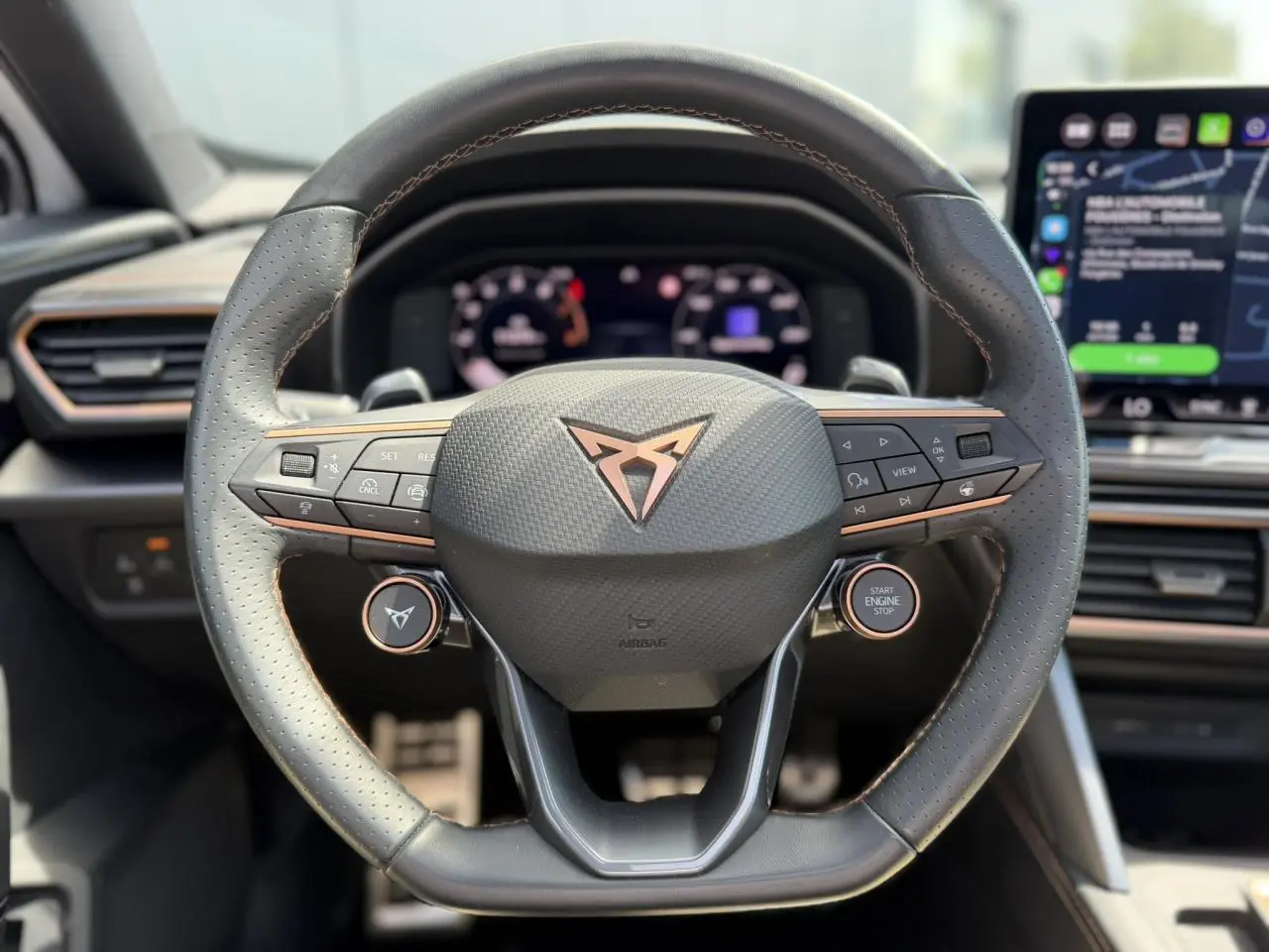 Volant Supersports CUPRA en cuir perforé avec logo cuivre, boutons satellites et tableau de bord numérique en arrière-plan.