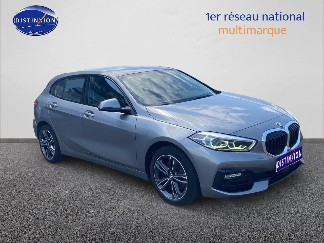 BMW Série 1 gris foncé vue 3/4 avant droit avec phares LED allumés et jantes alliage 17 pouces double rayons.