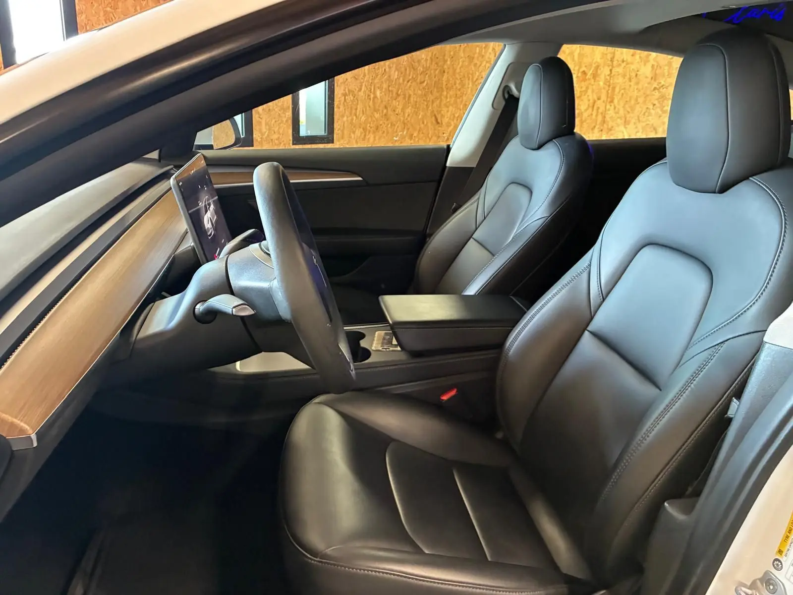 Vue intérieure côté conducteur de la Tesla Model 3 blanche 2023, sièges en cuir noir et tableau de bord avec écran tactile central.