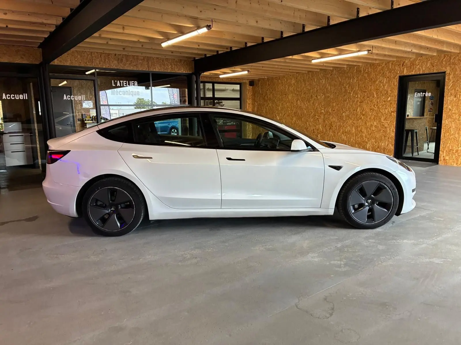 Vue de profil côté gauche d'une Tesla Model 3 blanche RWD 2023, avec jantes noires distinctives, dans un garage.
