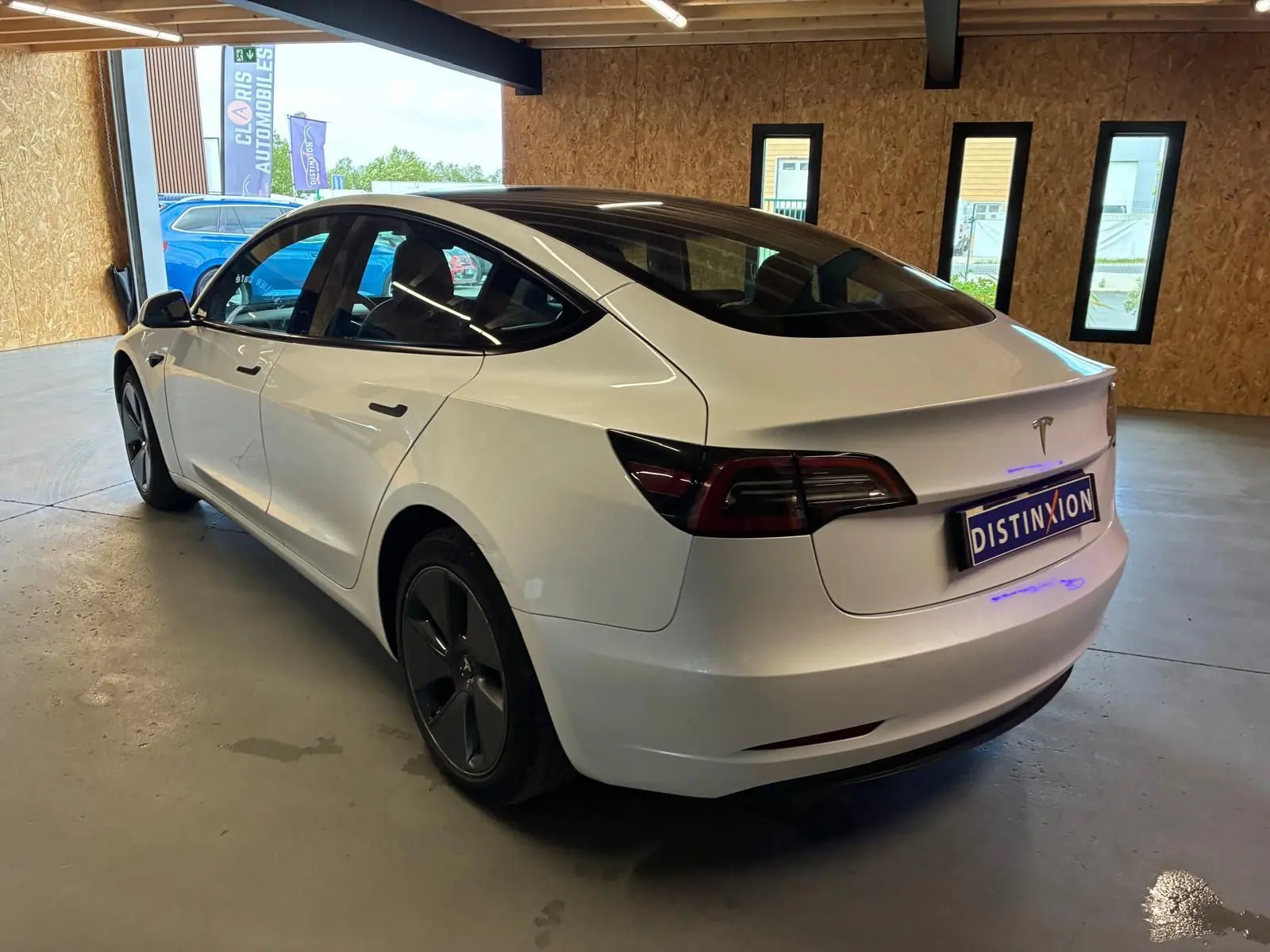 Tesla Model 3 blanc vue 3/4 arrière droit dans un garage, avec toit panoramique et jantes noires distinctives.