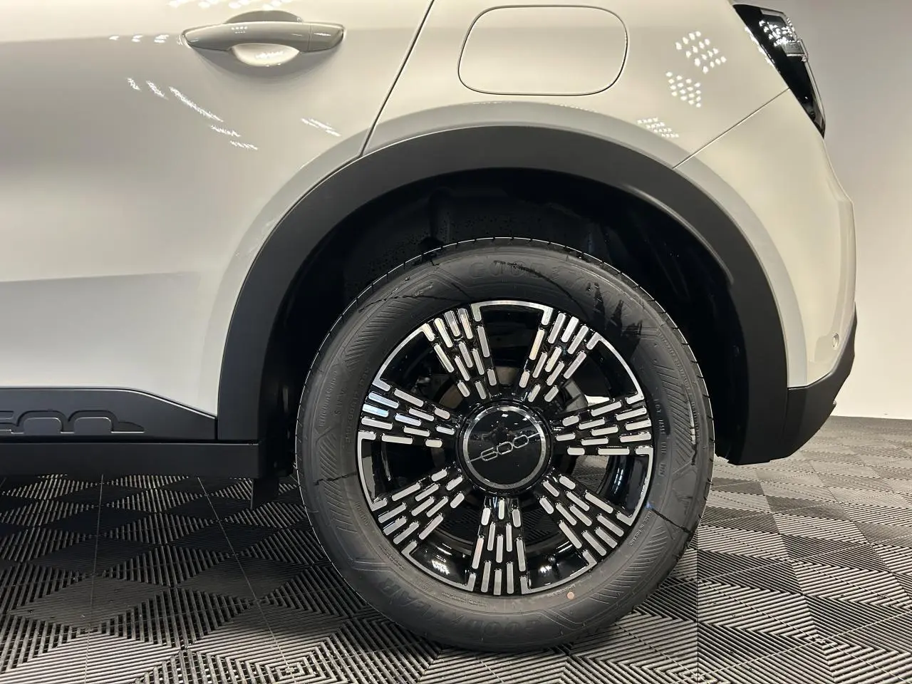 Gros plan sur la roue arrière droite noire et argentée du Fiat 600 blanc, avec passage de roue et partie de la carrosserie visibles.