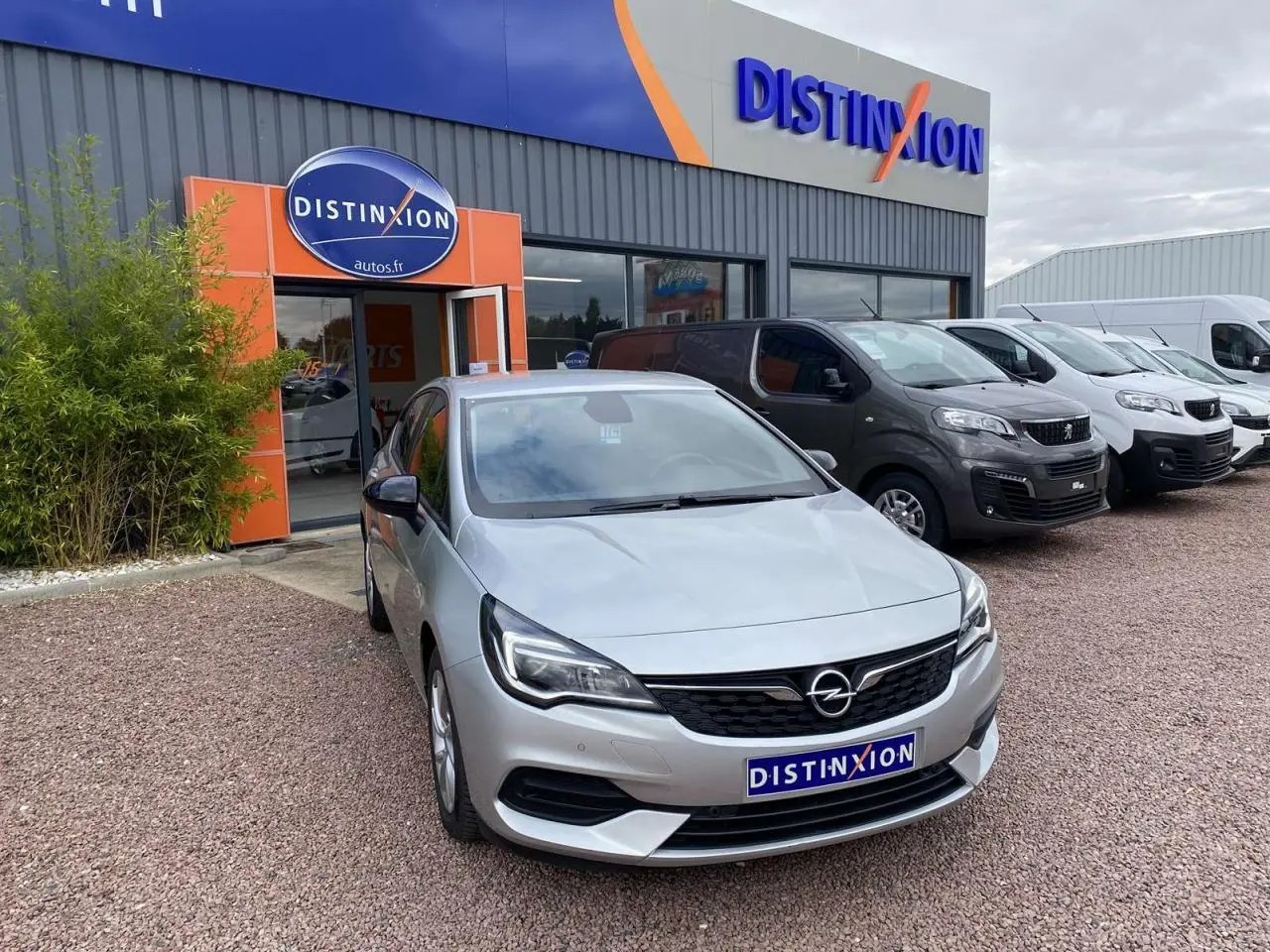 Opel Astra 1.2i Turbo gris argent vue 3/4 avant, avec calandre noire et phares LED, devant un garage Distinxion.
