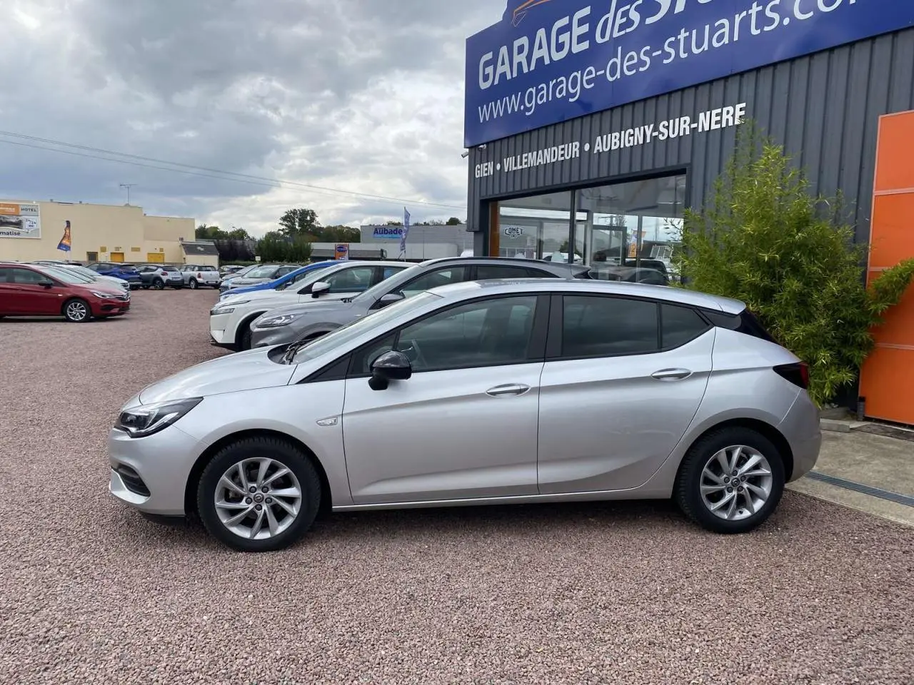 Profil côté gauche d'une Opel Astra 2021 Sovereign Silver avec jantes alliage 16 pouces, stationnée devant un garage.