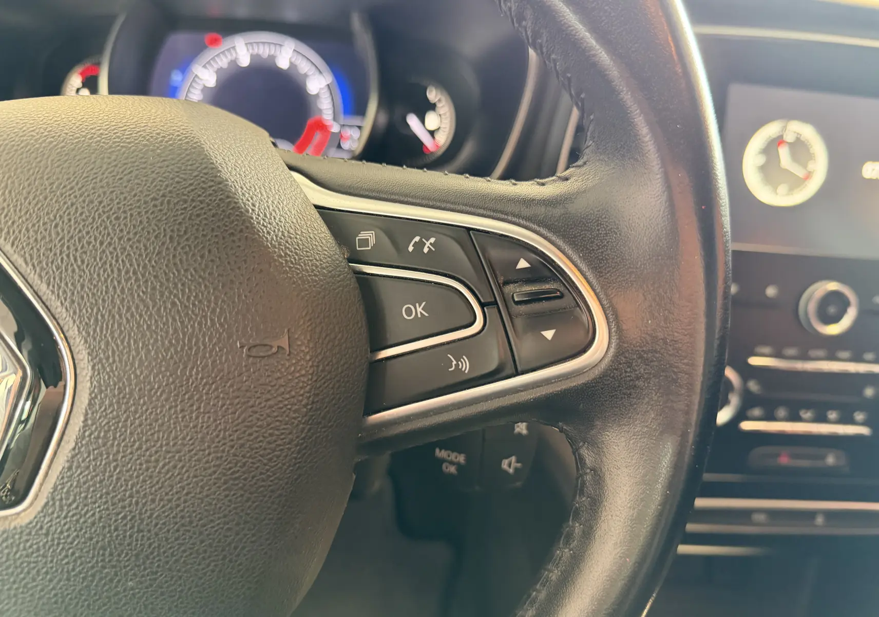Gros plan sur le volant en cuir noir côté droit de la Renault Mégane 1.3 TCE 115 Business avec commandes intégrées et tableau de bord lumineux en arrière-plan.