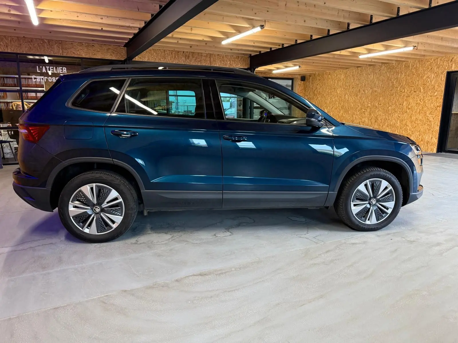 Vue de profil côté gauche d'un Skoda Karoq bleu 2023 avec jantes alu 17'' dans un garage en bois.
