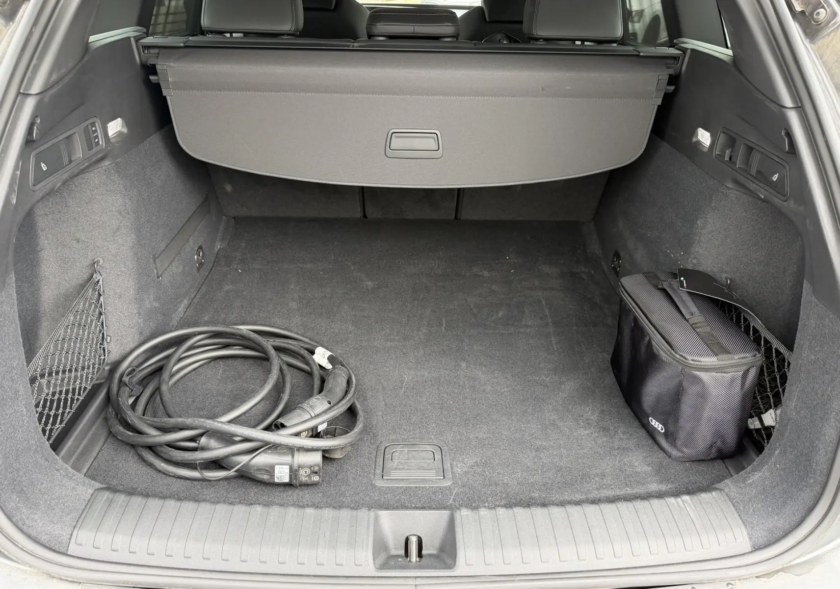 Coffre arrière ouvert de l’Audi Q6 e-tron gris, vue plongeante montrant câble de recharge et sac de rangement noir.