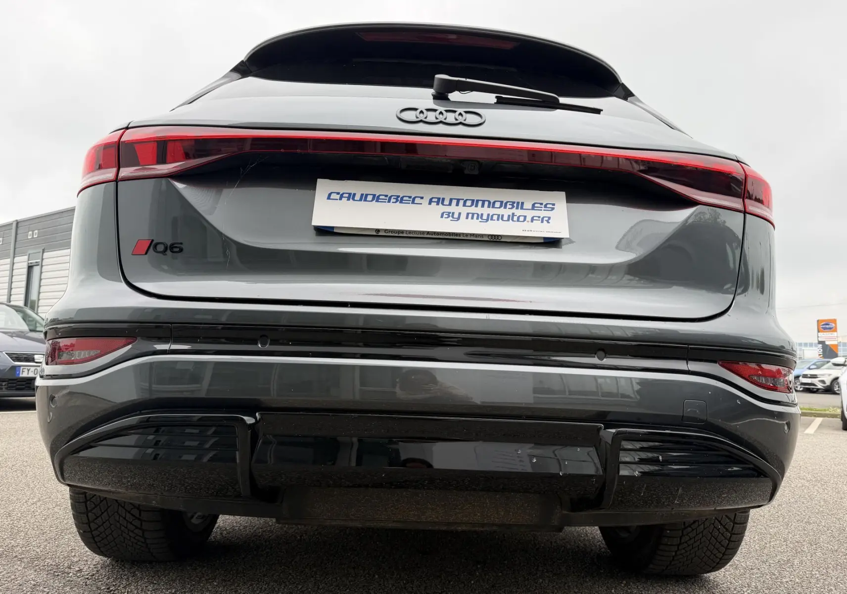 Vue arrière basse d’un Audi Q6 e-tron gris nacré avec feux arrière continus et logo Q6 rouge à gauche.