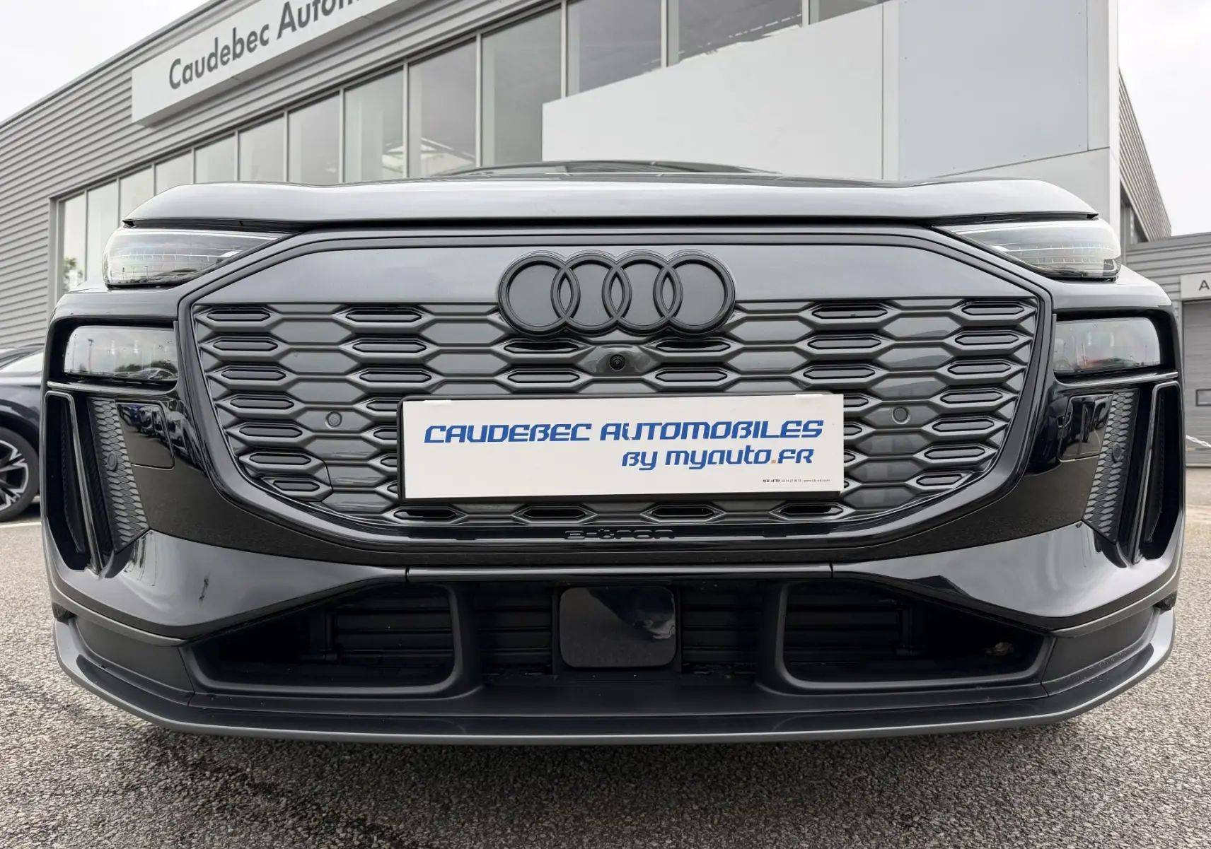 Vue frontale rapprochée de l'Audi Q6 e-tron gris nacré avec calandre hexagonale noire et logo Audi noir mat.