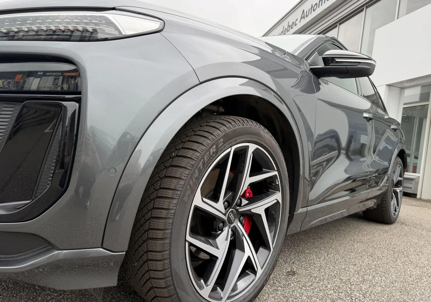 Vue 3/4 avant droit de l'Audi Q6 e-tron gris nacré mettant en valeur les jantes Audi Sport noires et étriers rouges.