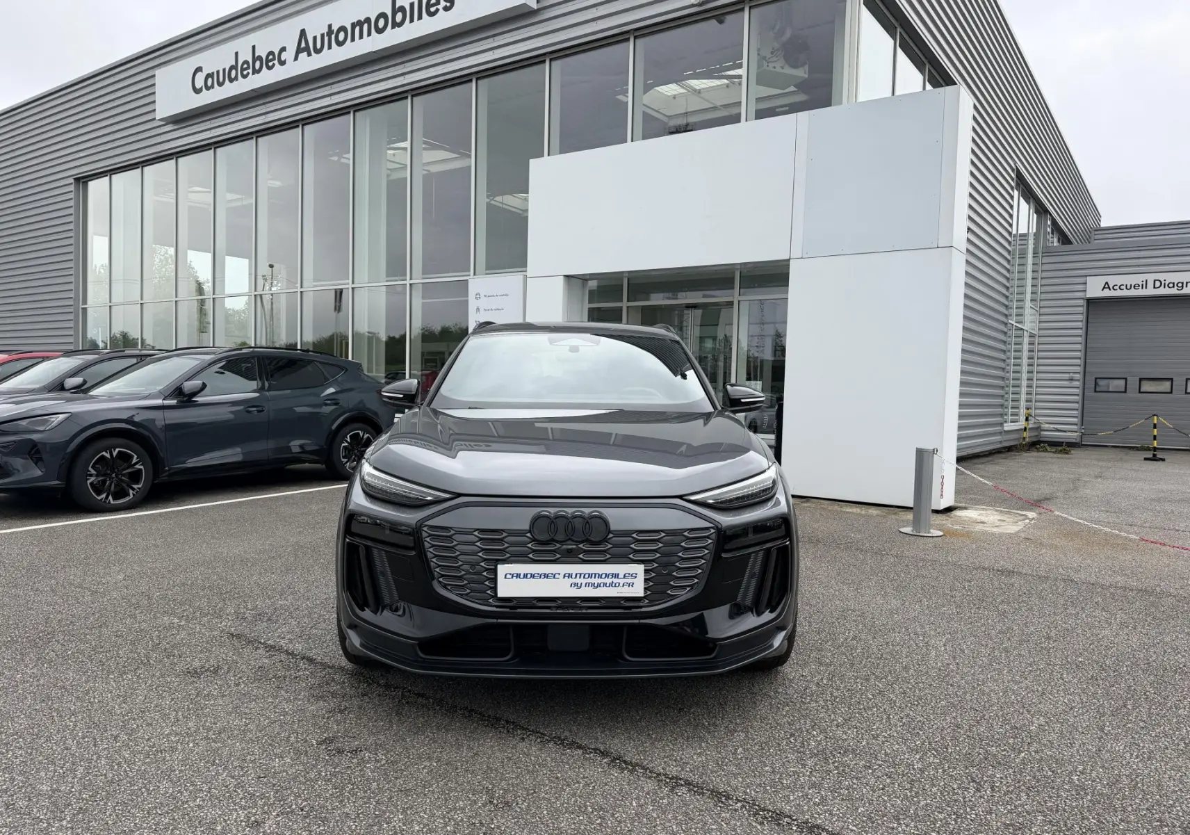 Vue avant d’un Audi Q6 e-tron gris nacré stationné devant un concessionnaire avec calandre et phares LED distinctifs.