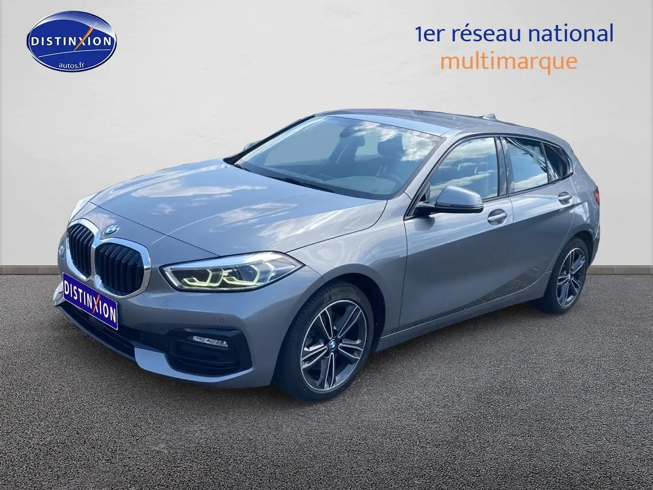 BMW Série 1 gris foncé vue 3/4 avant droit avec phares LED allumés et jantes alliage double rayons.