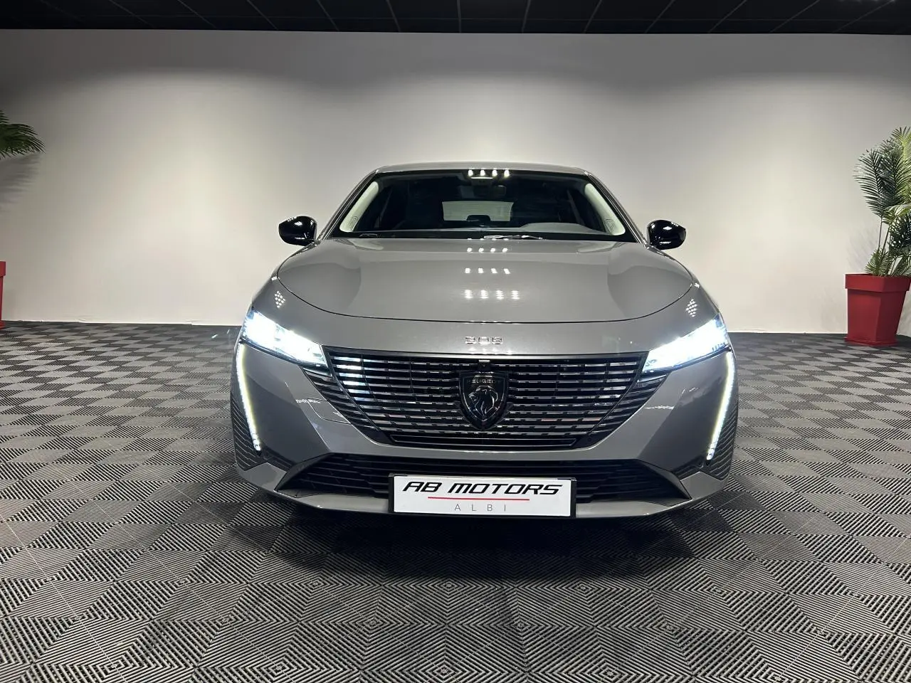 Peugeot 308 SW hybride gris Sélénium vue de face avec calandre noire et feux LED allumés dans un showroom.