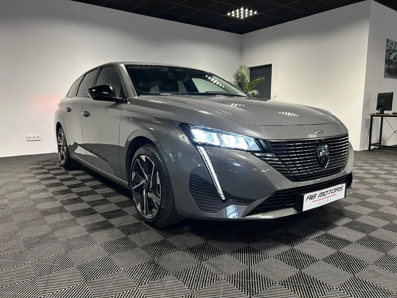 Peugeot 308 SW gris Sélénium vue 3/4 avant droit avec calandre noire et feux LED allumés en intérieur showroom.