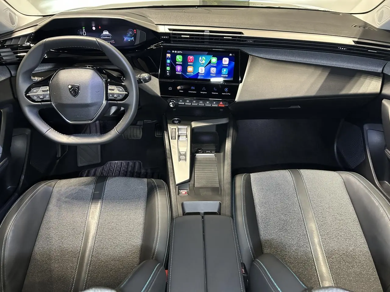Vue intérieure avant de la Peugeot 308 SW 2025, tableau de bord noir avec écran tactile et sièges bi-matière gris et noir.