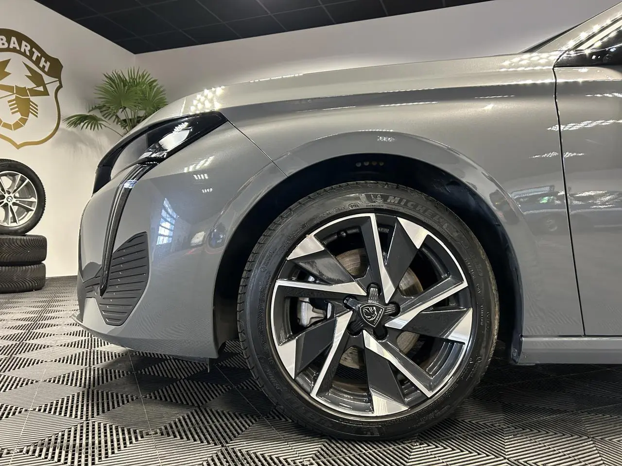 Gros plan sur la roue avant droite et l’aile avant gris Sélénium d’une Peugeot 308 SW hybride 2025 en showroom.