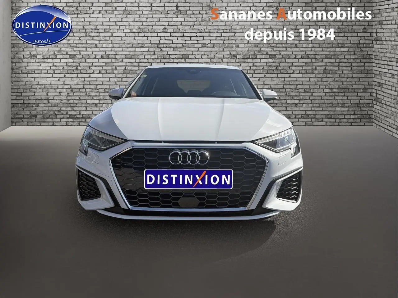 Vue frontale d'une Audi A3 Sportback blanche 2023 avec calandre noire et projecteurs LED, sur fond mural en briques.