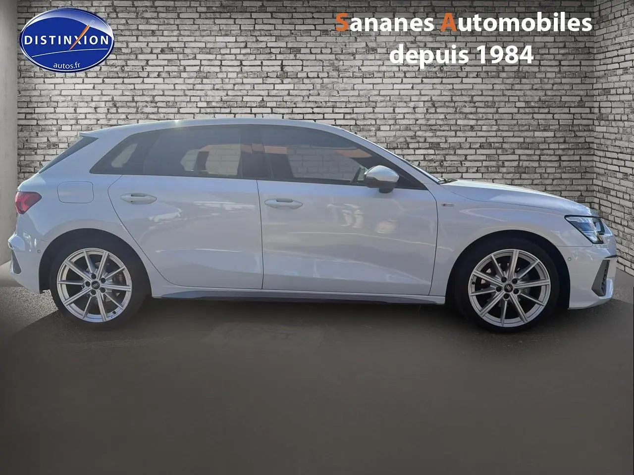 Profil droit d'une Audi A3 Sportback blanche 2023 avec jantes alliage et détails S line visibles sur fond de mur en briques.