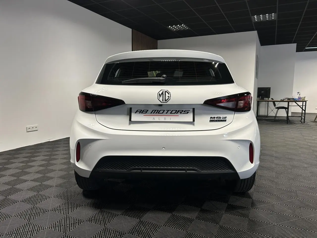 Vue arrière d'une MG3 1.5 Hybrid+ blanche 2025 en intérieur, avec feux arrière fumés et logo MG bien visible.