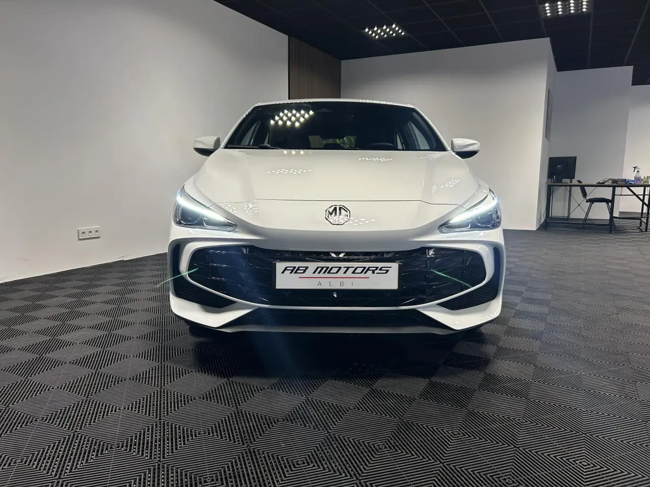 Vue frontale d'une MG3 blanche 2025 avec phares LED allumés dans un showroom moderne.