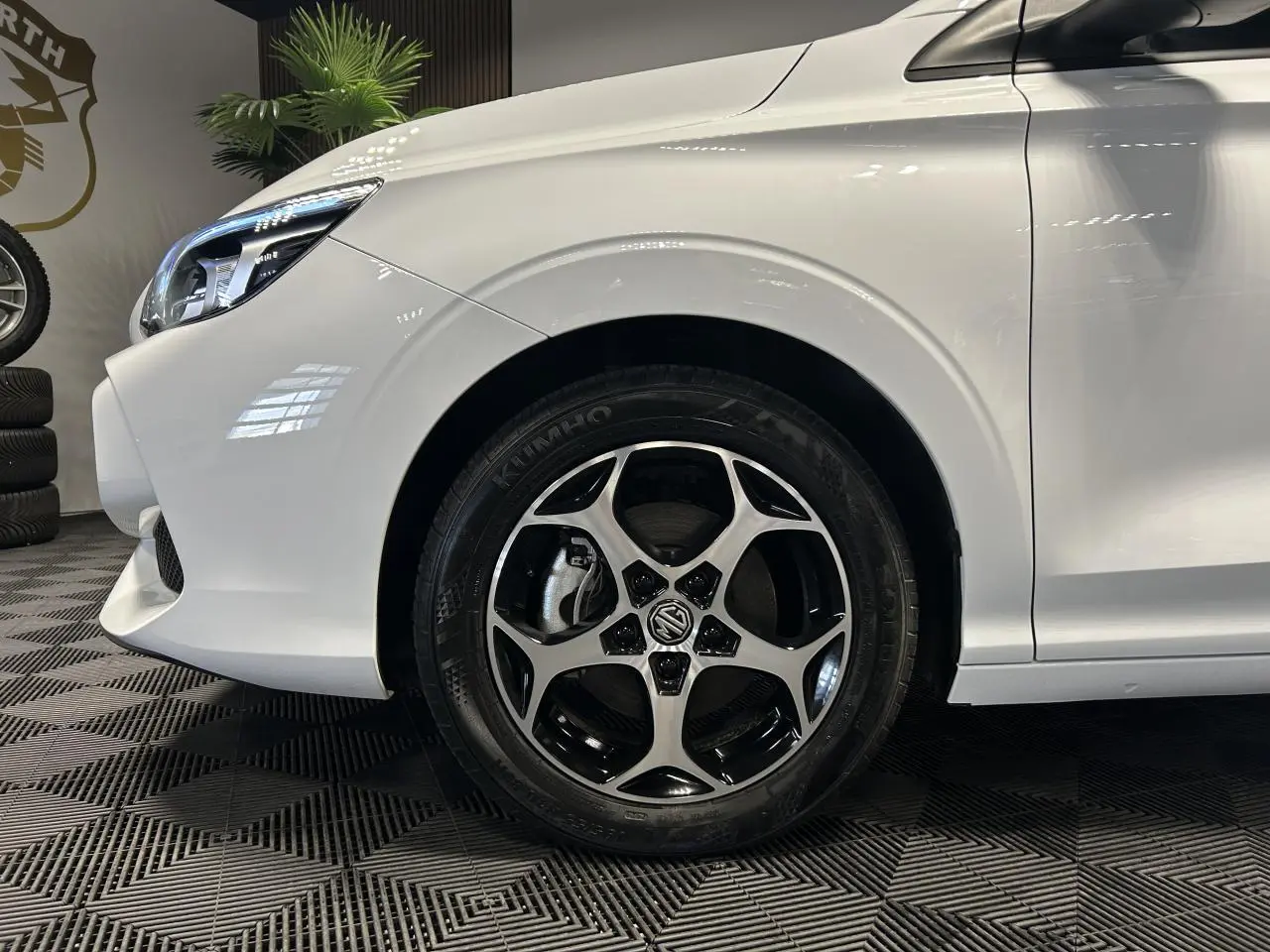 Gros plan sur la roue avant gauche et le phare d’un MG3 blanc 2025 dans un intérieur showroom.