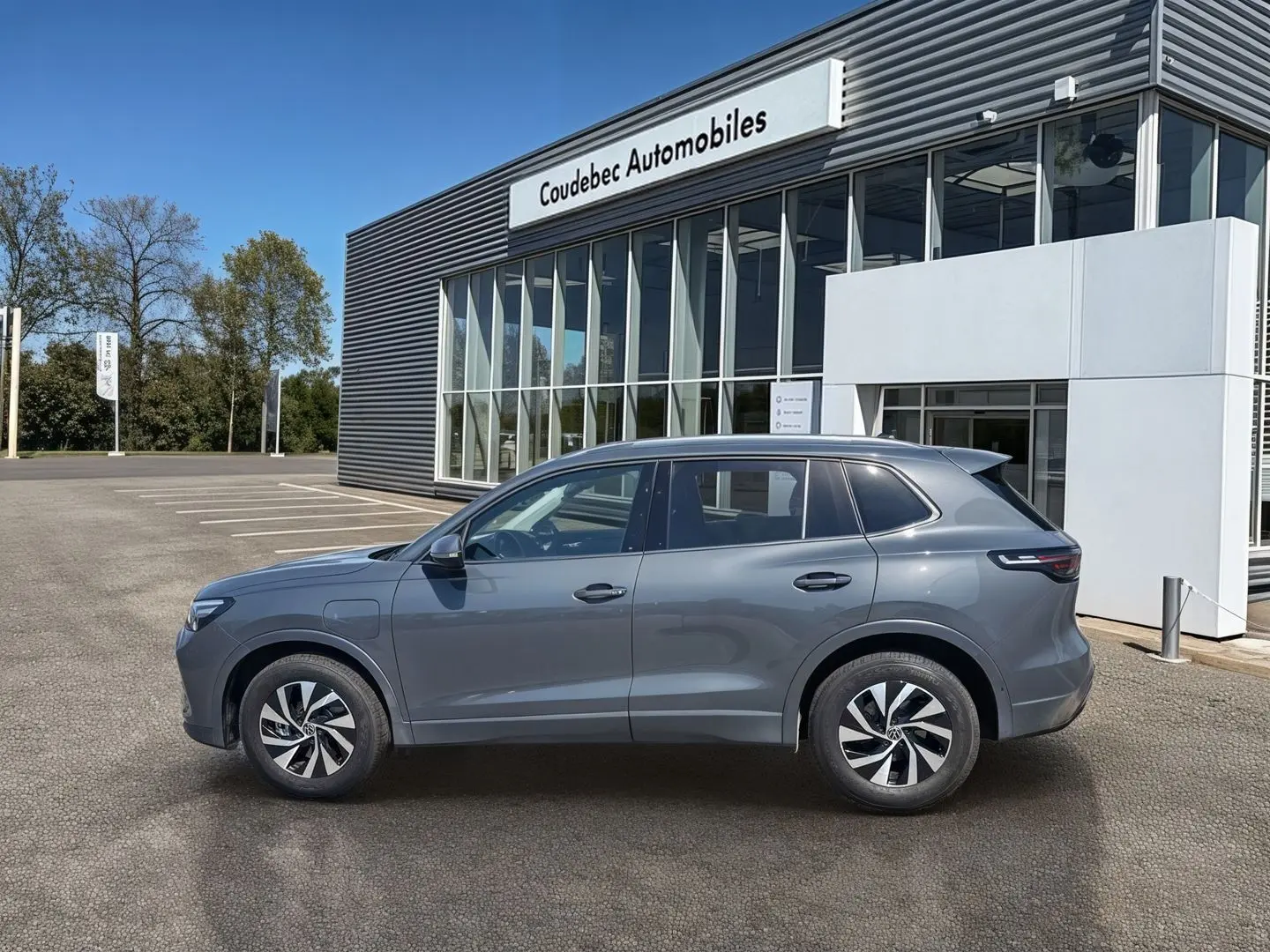 Vue latérale droite d'un Volkswagen Tiguan 1.5 eHybrid gris, avec jantes alliage Venezia 17 pouces, devant un bâtiment commercial.