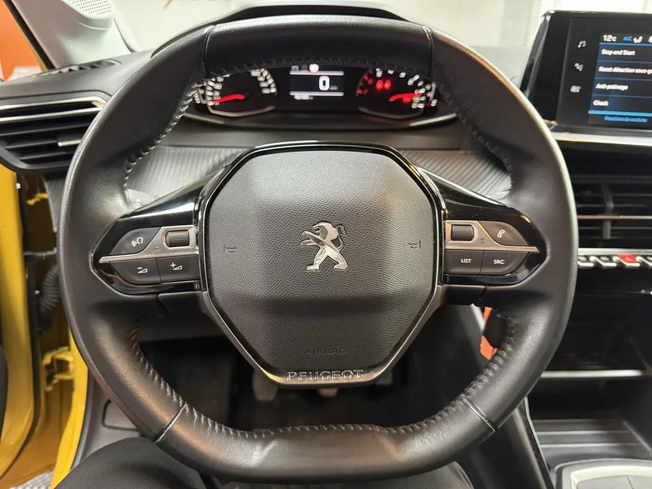 Vue centrée sur le volant cuir noir de la Peugeot 208 2020 avec tableau de bord et écran tactile visibles, portière jaune ouverte.
