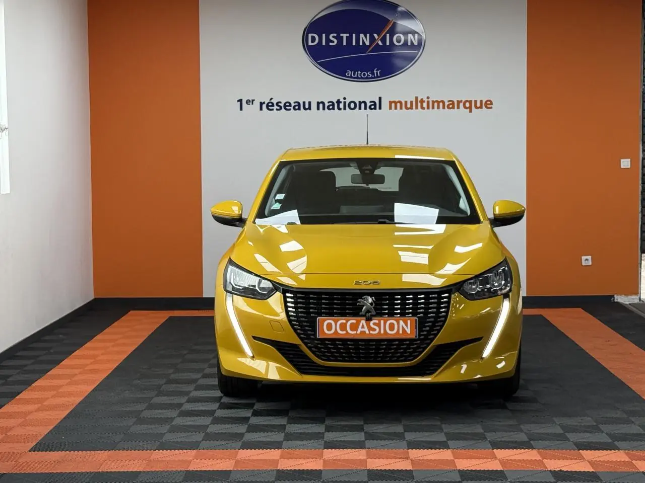 Vue frontale d'une Peugeot 208 jaune clair avec calandre noire et plaque "OCCASION" dans un showroom.