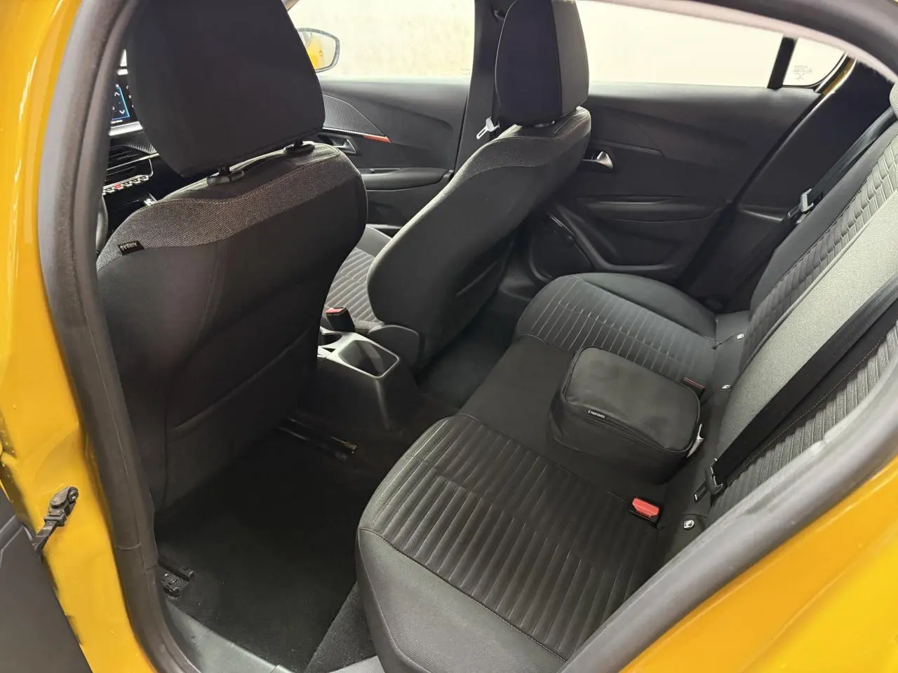 Vue intérieure arrière droite d'une Peugeot 208 jaune clair, montrant les sièges noirs et un sac posé sur la banquette.