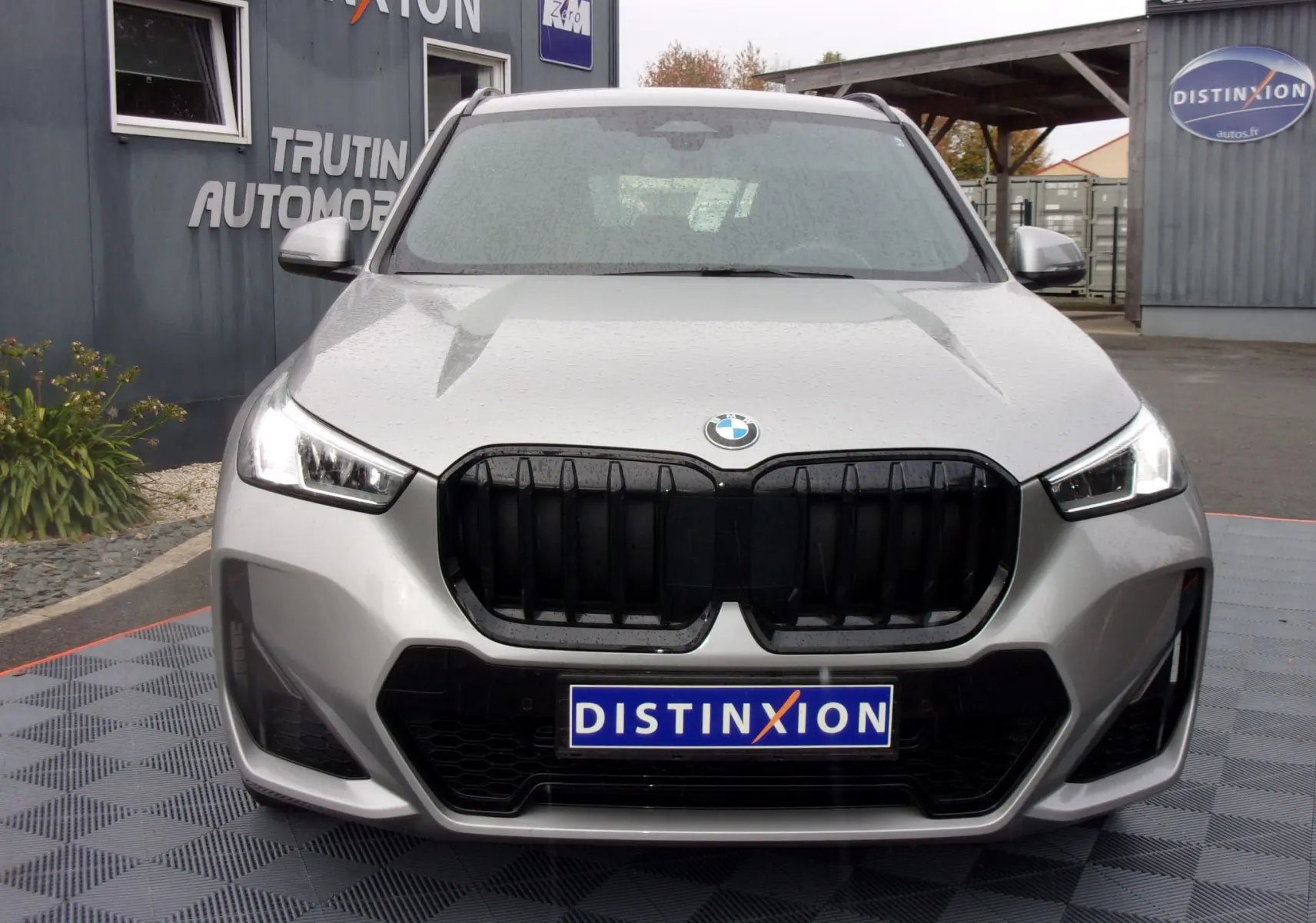 Vue frontale d'un BMW X1 2024 argent métallisé avec calandre noire et phares LED allumés sous la pluie.