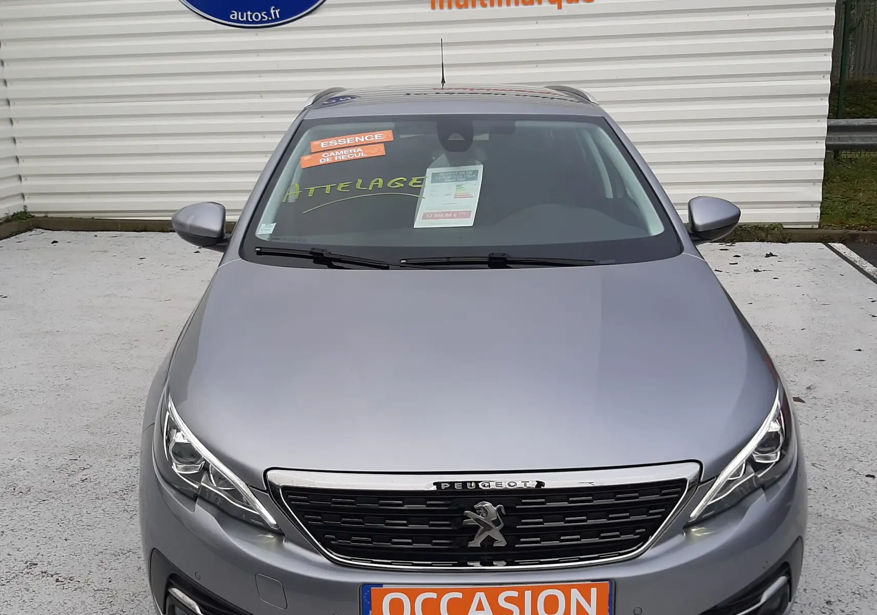Vue avant d'une Peugeot 308 SW gris Artense avec plaque "OCCASION" et autocollants sur pare-brise dans un parking.