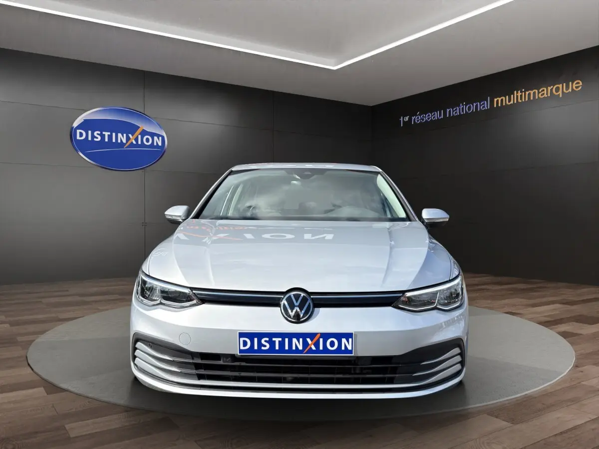 Vue frontale d'une Volkswagen Golf gris clair 2022 avec logo VW et plaque Distinxion visible en showroom.