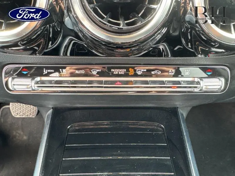 Gros plan sur la console centrale noire brillante d'une Mercedes GLA 250 e bleu denim, montrant les commandes climatisation et sécurité.