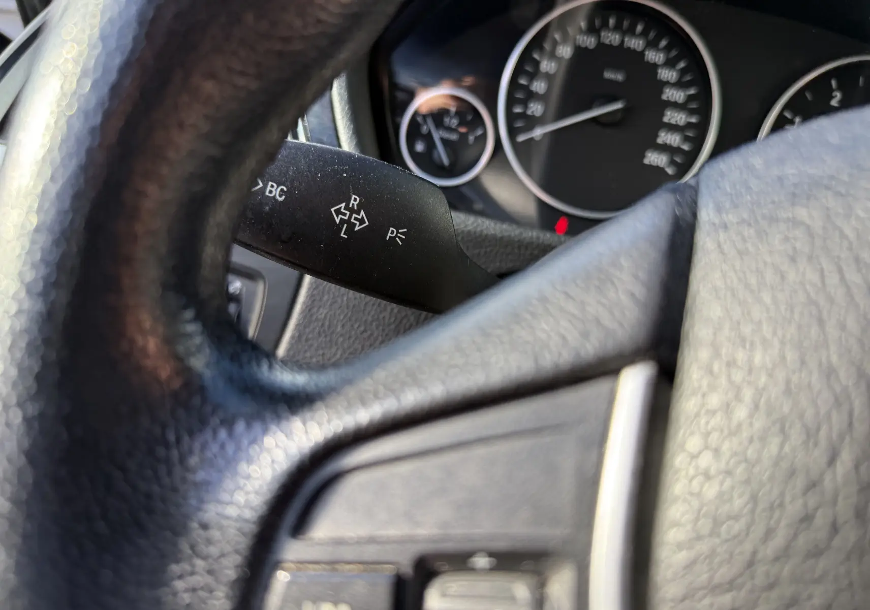 Gros plan sur le volant noir et le commodo gauche de la BMW Série 3 320D gris, avec compteurs en arrière-plan flous.