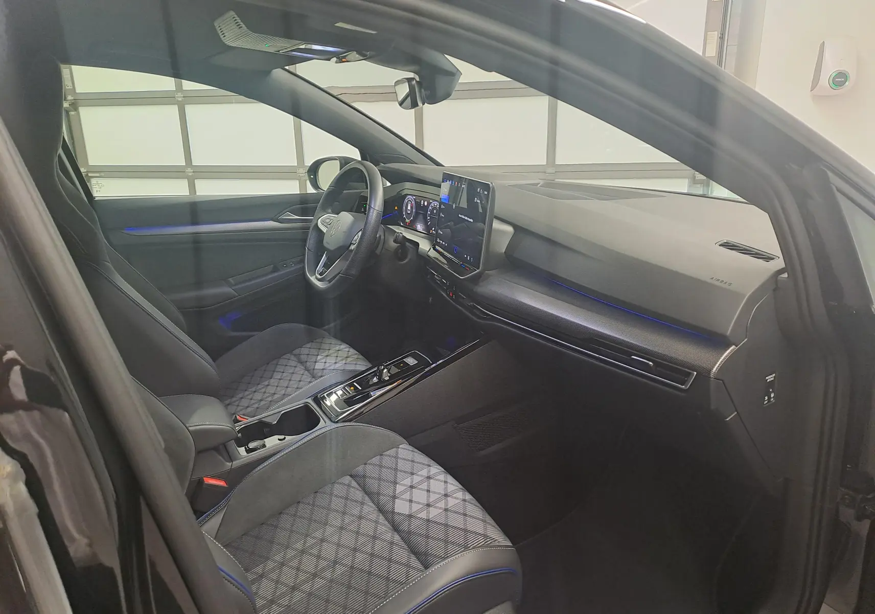 Vue intérieure côté conducteur de la Volkswagen Golf 2.0 TDI 150 DSG7 R Line 2024 avec sièges tissu à motif et éclairage d'ambiance bleu.